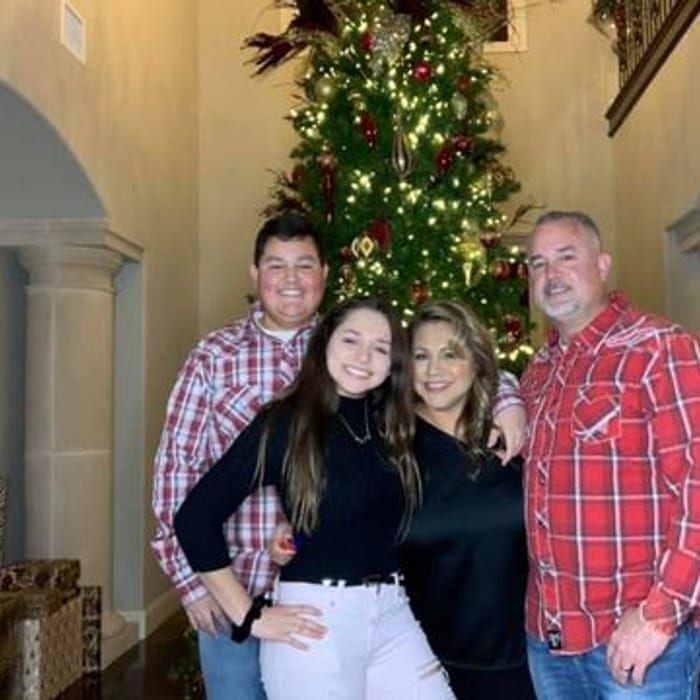 Family posing in front of a decorated Christmas tree. People are smiling, wearing casual clothes. Brightly lit interior.