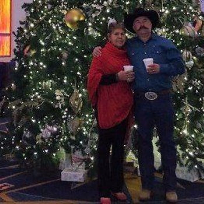 Couple posing in front of a Christmas tree with drinks; woman in red poncho, man in cowboy hat.